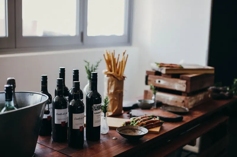 Various bottles of alcoholic drinks placed on table with grissini sticks and wooden boxes near green branches in vase in room